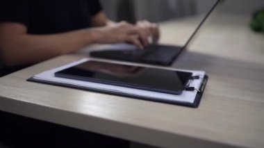 Tablet yakınlardaki not defterinde dururken, kimliği belirsiz kişi temiz bir masada dizüstü bilgisayarda yazıyor. Çalışma alanı minimalist tasarım elementlerine sahiptir. Mavi arka planda dizüstü bilgisayarda yazan kadın elleri.