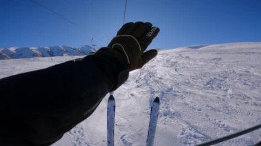 Kayakçı teleferiğe biniyor ve kayak eldivenleriyle yüzünü güneş ışığından koruyor. Kış manzarasının tadını çıkaran kişi, tatilde sandalyede gezmekten hoşlanır..