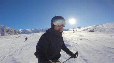 Koyu renkli kayakçı kostümlü koruyucu kasklı ve refleks gözlüklü kayakçıyı kapatın. Güneşli kış günlerinde açık mavi gökyüzünün altında kar altında kayarak ilerleyin..
