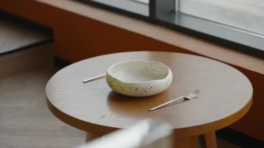Çağdaş restoranda seramik beyaz kâse ve iki parıldayan çatal bıçak ile hazırlanmış minimalist masa. Kafede masa ayarı. Dekoratif tabak, çatal ve kaşık ahşap yuvarlak masa..