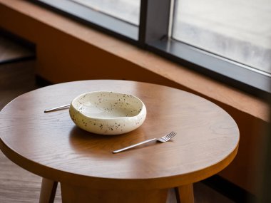 El yapımı seramik kâse büyük bir pencereden gelen doğal ışıkla aydınlatılmış yuvarlak ahşap bir masanın üzerinde duruyor. İki çatal, rahat bir iç mekandaki minimalist yemek düzenini tamamlar..