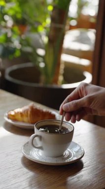 Erkek eli, sıcak ve davetkar bir kafede bir tabak yumuşak hamur işiyle bir fincan koyu kahveyi karıştırır. Güneşli bir günde kafenin rahatlatıcı atmosferi. Güneş ışığıyla dolu restoranda kahvaltı..