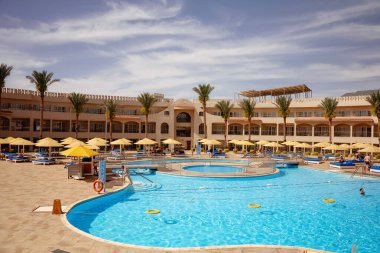 Mısır, Sharm El Sheikh - 18 Mart 2025: Palmiye ağaçları ve sarı şemsiyelerle çevrili büyük yüzme havuzlu canlı bir tatil köyü. Mavi su havuzlu bir daire ve şemsiyeli şezlonglar.