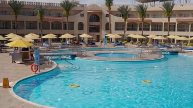 Mısır, Sharm El Sheikh - 18 Mart 2025: Lüks otel temiz mavi suyla yüzme havuzunun arkasındaki modern apartman dairesi, güneş altında şezlongları ve şemsiyeleri davet ediyor.