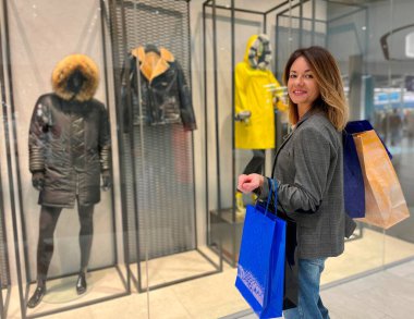 Alışveriş merkezinde alışveriş torbalarıyla duran ve alışveriş yaparken kameraya gülümseyen mutlu yetişkin bir kadın. Neşeli beyaz kadın, kışlık palto giymiş, vitrinin yanında duruyor. Satış konsepti