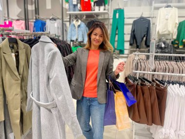 Alışveriş merkezinde alışveriş yapan, çantalarıyla ceketini gösteren kameraya bakan mutlu, beyaz bir kadın. Moda mağazasında kıyafet seçen bir kadın. Alışveriş koliği, satın alma kavramı