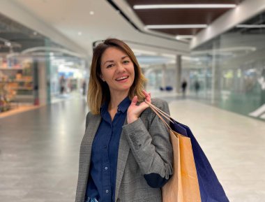 Mutlu yüzlü, orta yaşlı beyaz bir kadın alışveriş merkezinde kameraya bakıp gülümsüyor. Güzel, mutlu, kağıt torbalı, alışverişten sonra mutlu bir kadın. Pozitif duygular