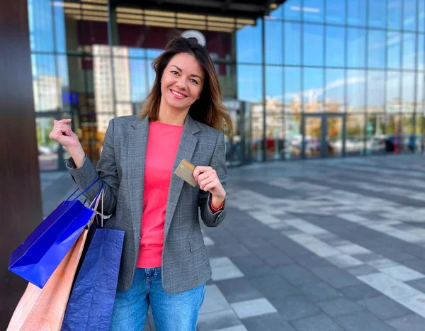 Gülümseyen güzel bir kadın alışveriş merkezinin yakınındaki kameraya bakıyor. Elinde hediye ve kredi kartı dolu çantalar var. Tatil alışverişi. Noel zamanı