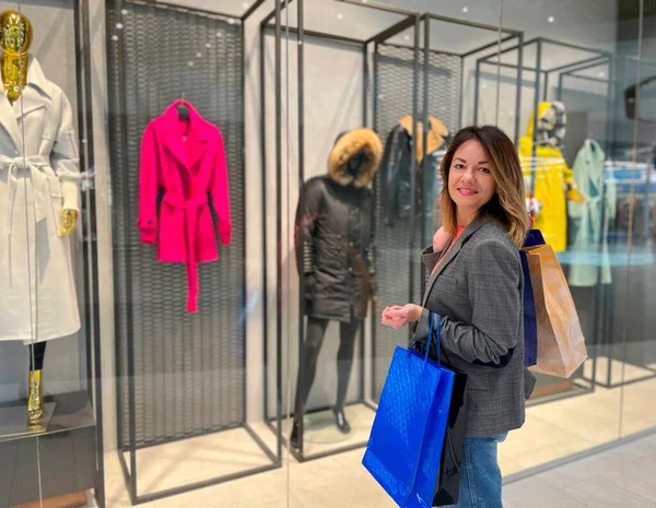 Alışveriş merkezinde poşetlerle durup alışveriş yaparken kameraya gülümseyen yetişkin beyaz bir kadın. Moda kıyafetleriyle vitrinin yanındaki neşeli kadın. Alışverişkolik kavram
