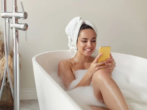 Close up of Caucasian beautiful woman relaxing in hot bath with foam and bubbles texting on smartphone. Cheerful young female taking bath and using mobile phone. Leisure time. Bathing concept