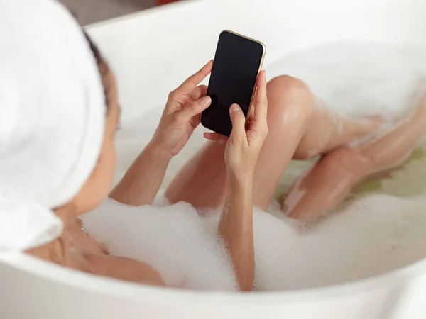 浴槽の女性の手でスマートフォンの画面を閉じます 裸の女性の肩越しに浴槽に横たわってスマートフォンに入力します 女性はバスに乗って携帯電話でオンラインで閲覧 — ストック写真