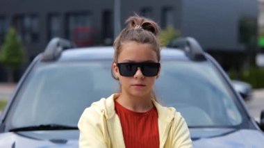 Beyaz havalı genç kız caddede arabanın önünde durmuş sakız çiğneyen kameraya bakıyor. Kapatın. Ciddi yüz ifadesi olan şık bir genç kız. Açık hava, şehir yaşam tarzı. Yavaş çekim