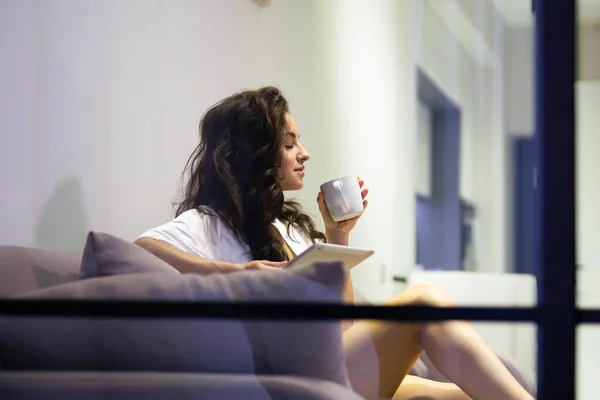 Beautiful Caucasian young relaxed woman in robe resting on sofa in modern living room drinking coffee holding cup in hands. Attractive pretty female indoors drinks tea. spying through the glass