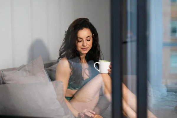 Spying on beautiful Caucasian young relaxed woman in lingerie resting on sofa in modern living room drinking coffee and eating croissant. Pretty female in underwear having breakfast. Keeking concept