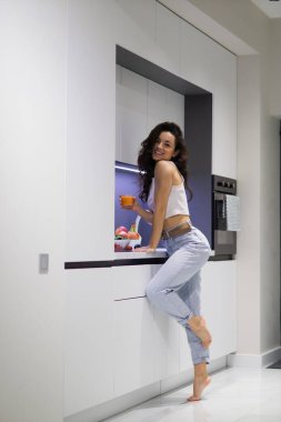 Cheerful young Caucasian attractive female standing in modern kitchen at home holding glass of fresh orange juice in hand and looking at camera smiling. Breakfast time, vertical shot. Indoor concept.