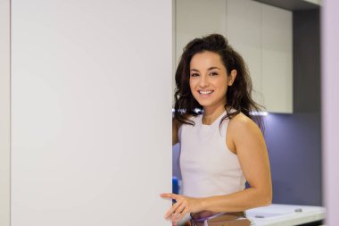 Close up portrait of young cheerful brunette woman smiling and looking at camera while standing in kitchen opening door of refrigerator. Happy positive casual housewife at home indoors