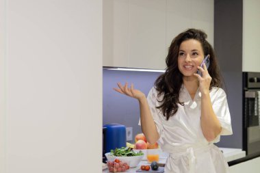 Young attractive beautiful happy smiling woman in white robe standing in modern kitchen and talking on smartphone. Pretty female at home in morning calling on cellphone in apartment. Portrait shot