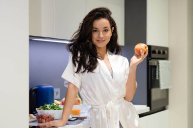 Happy young attractive brunette female housewife holding apple in hands smiling while looking at camera standing in kitchen. Pretty Caucasian female in robe at home. Healthy lifestyle concept
