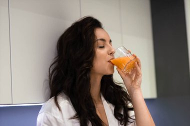 Close up of attractive young Caucasian female with dark long hair in white robe in modern kitchen having breakfast holding glass of orange juice. Beautiful young woman drinking fresh juice at home