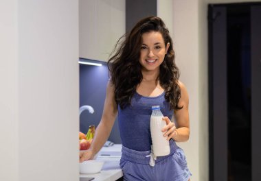 Portrait of happy smiling young Caucasian dark-haired woman in blue pyjamas standing in modern kitchen with bottle of milk in hands and looking at camera. Beautiful joyful female preparing breakfast