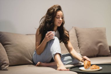 Beautiful attractive lady in casual clothes sitting on couch indoors holding in hands cup of hot drink and taking tasty croissant from plate. Caucasian woman having breakfast on sofa at cozy home