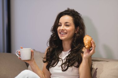 Happy smiling young attractive female resting at home holding in hands cup of tea and croissant. Close up concept. Joyful beautiful brunette lady with happy face enjoys coffee with pastry indoor