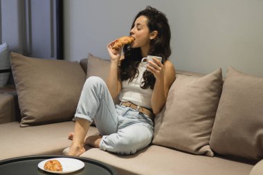 Attractive stylish woman sitting on sofa in modern apartment drinking coffee and eating croissant. Caucasian beautiful concented brunette female has breakfast at cozy home. Food lover concept