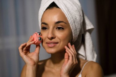 Close up of Caucasian young attractive cheerful woman with towel on head using microcurrent facial toning device at home. Beauty procedure. Beauty device. Beautiful happy female using skincare device
