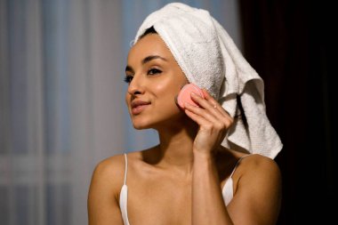 Close up of pretty young joyful smiling woman with towel on head using modern facial toning device indoor at home. Beauty procedure. Beautiful happy female using skincare device. Beauty device concept