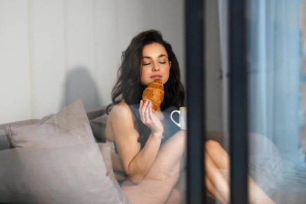 Caucasian young pretty relaxed woman in sexy green lingerie sitting on sofa in modern living room drinking coffee and eating croissant. Brunette female in underwear having breakfast. Spying concept
