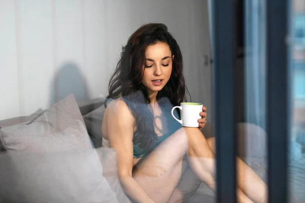 Spying through the glass on attractive sexy young woman in lingerie resting on couch at home holding in hands cup of hot drink. Close up. Pretty female in seductive underwear in living room