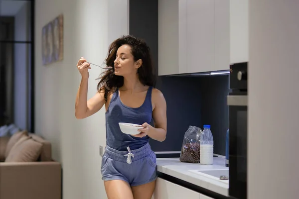 Adult young beautiful Caucasian woman indoors at home eating breakfast. Young pretty brunette female with long hair enjoying cereal eating from spoon standing in the kitchen in morning