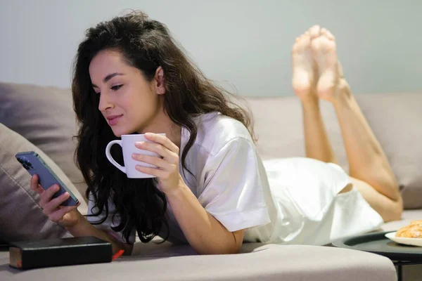 Beautiful young woman in white robe lying on couch in living room alone tapping on smartphone holding cup of coffee. Caucasian brunette female resting on sofa at home using mobile phone. Close up
