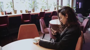 Uzun siyah saçlı, boş bir restoranın içinde cappuccino içen güzel beyaz kadın odanın ortasında yuvarlak masada oturuyor ve akıllı telefonunun ekranına bakıyor..