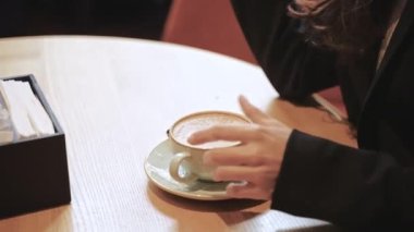Uzun siyah saçlı, beyaz kahve fincanından kapuçino içen beyaz bir kız yuvarlak ahşap bir masada oturuyor ve akıllı telefonunun ekranına bakıyor..