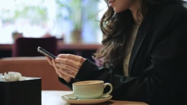Beautiful Caucasian woman with long brown hair sitting inside a good modern restaurant, drinking coffee, smiling, and typing on her smartphone. Close-up, slow motion, face, smile, eyes.