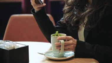 Yakın çekim, yavaş çekim. Modern kafenin içinde bir sürü yeşil bitkiyle oturan, naneli sıcak çay içen, gülümseyen ve akıllı telefonunu kaydıran güzel beyaz kadın..