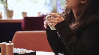 Close-up, slow motion, fingers, cup. Beautiful Caucasian girl sitting inside of modern cafe with a lot of green plants, drinking hot tea with mint with pleasure and touching her long brown hair.