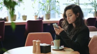 Yakın çekim, yavaş çekim. Kafkasyalı güzel iş kadını bir sürü yeşil bitkiyle modern restoranın içinde oturuyor, naneli sıcak çay içiyor ve telefonuna kayıyor ve daktilo çekiyor..