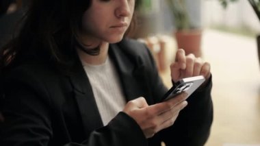 Esmer, uzun saçlı, modern bir restoranda oturan, kaydıran ve akıllı telefonuyla yazan beyaz bir kadın. Kız blogcu uzaktan çalışıyor, içerik yaratıyor ve ciddi ve yorgun görünüyor.