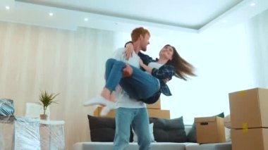 Young joyful married couple feeling happy together in new apartment. Low angle view. Loving young husband spinning his beloved beautiful wife on hands in new house on moving day. Own place to live