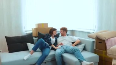 Camera approaching Caucasian joyful male and pretty female smiling jumping on a couch in light living room with many cardboard boxes. Boyfriend and girlfriend moving in together. Real time. Close up