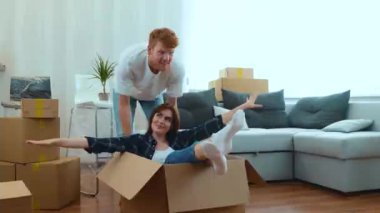Happy young couple having fun in new bought apartment. Funny husband and wife playing on moving day. Excited adult man pushing cardboard box with his girlfriend sitting inside. Relocate in new home