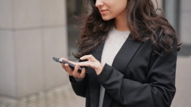 Close up, slow motion. Young businesswoman standing on the street next to modern grey building, looking at her phone, smiling, and touching her hair. Outdoors, business, and office buildings.