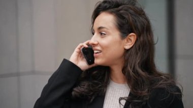 Young girl walking on streets next to grey modern buildings, talking by phone, and drinking hot drink from paper cup to go. Woman is smiling and looks happy. Outdoors, slow motion.