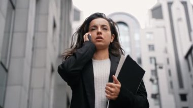 Businesswoman walking on the street and having conversation by phone. Young Caucasian girl talking by phone, carrying laptop, and files in her hand, walking outdoors around modern office buildings.