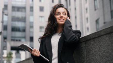 Young Caucasian business lady talking by phone standing outdoors around grey contemporary office buildings. Girl has laptop, and files in her hand and standing on the stairs, smiling, looking around.