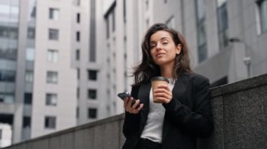 Caucasian beautiful female standing outdoors around grey modern buildings, working on her phone, smiling, looking around, and drinking drink from paper cup to go. Close up, slow motion.