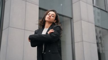 Back view, close up, slow motion. Woman with long dark hair and black suit turns to the camera, smiling, and putting her hands on her chest. Outdoors, grey office modern buildings, city center.