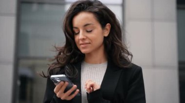 Young girl wearing office style jacket standing downtown. Caucasian businesswoman standing next to office buildings, smiling, looking around, touching her dark long hair, scrolling her smartphone.